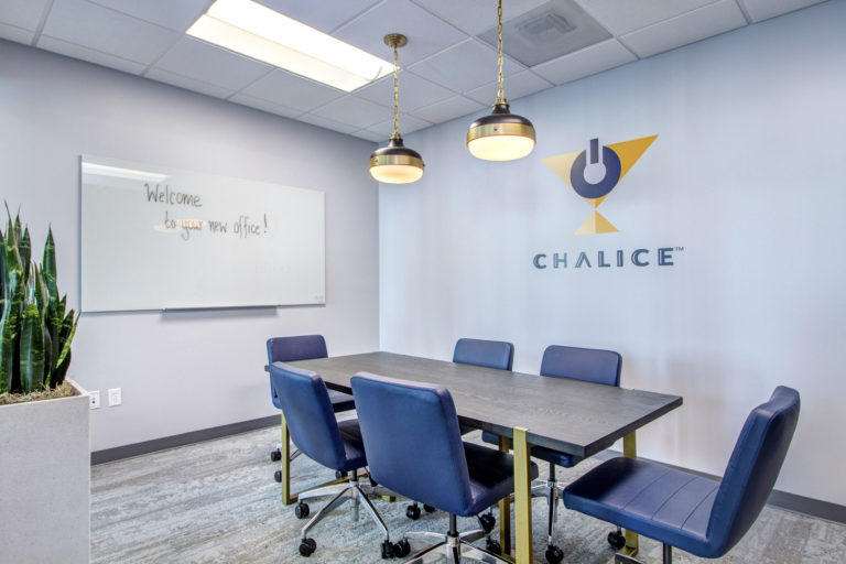 A conference room with 6 blue seats, a small conference table, and a logo on the back wall that reads "Chalice"