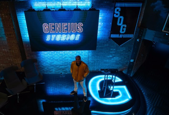 Billy Gene standing on a black stage with a bright blue light up G on the stage, and a screen behind Billy that says "Geneius Studios"
